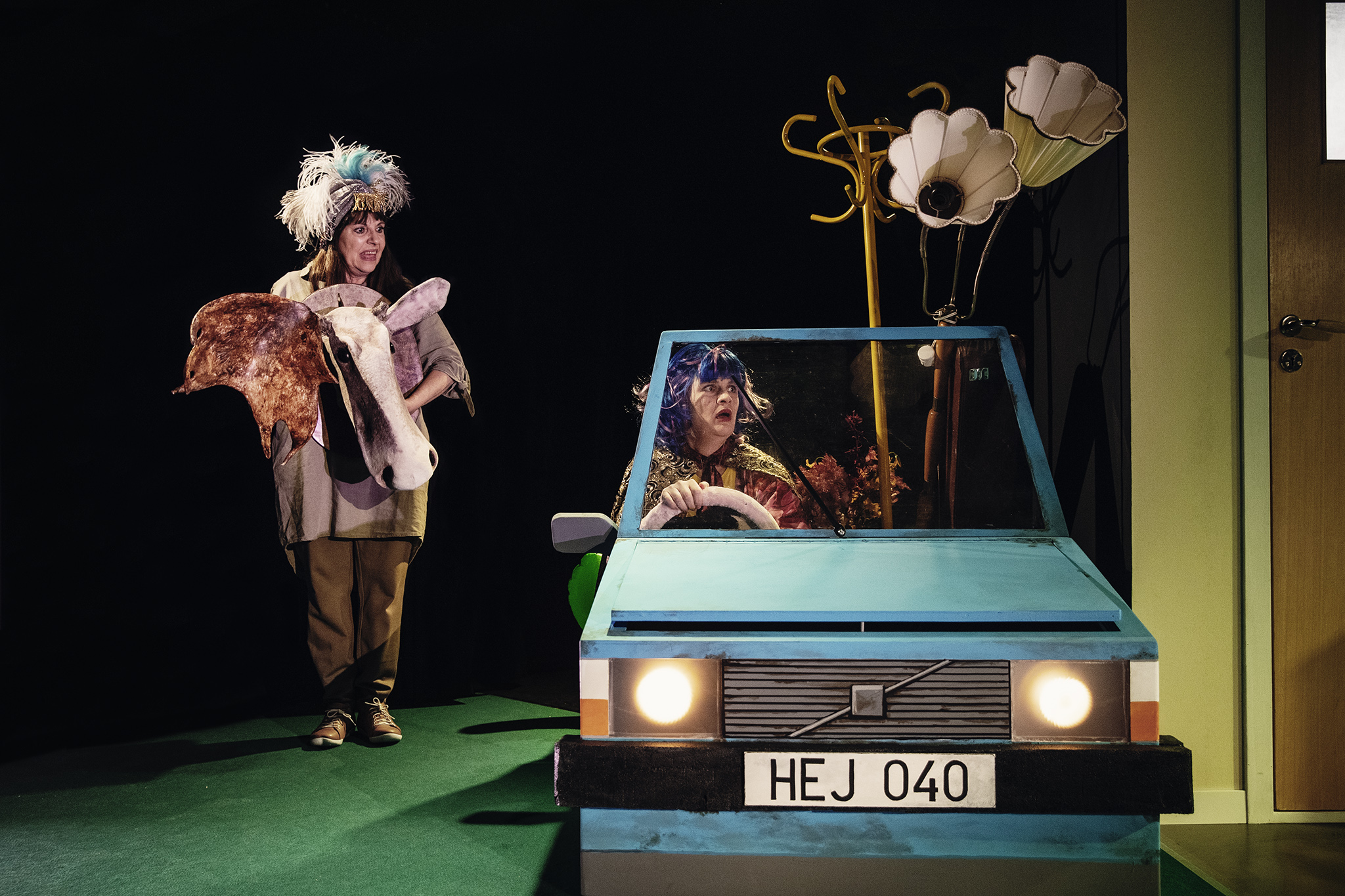 Foto från föreställningen på Jag (Ann Katrin Andréasson) som står bredvid Bettans bil med ett uppstoppat älghuvud i famnen. Bettan (Josefin Larsson) sitter i sin bil till höger.